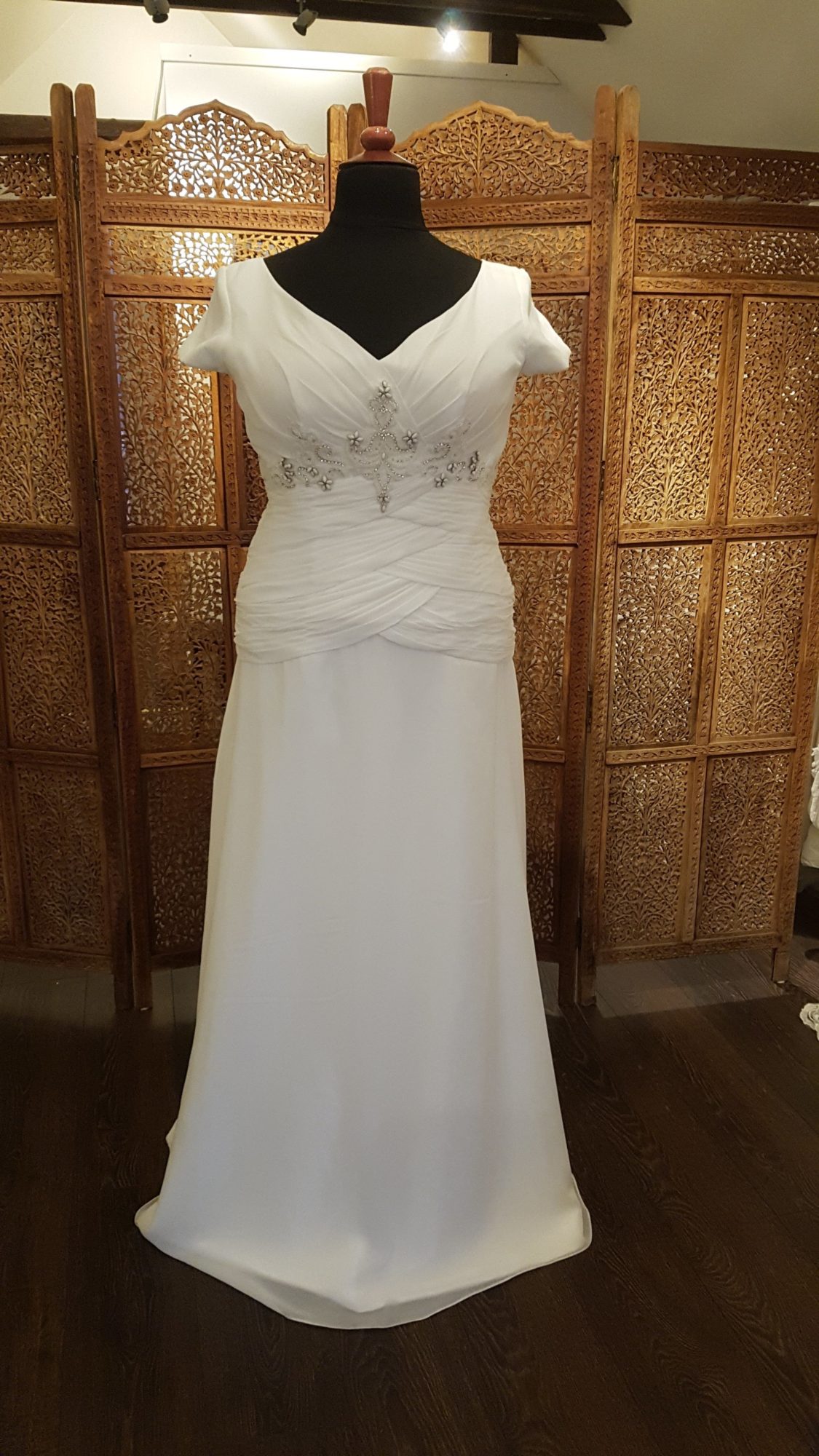 Smuk og enkel brudekjole i chiffon med en slank silhuet. Kjolen har draperinger omkring taljen, perlepynt under barmen og et lille ærme.