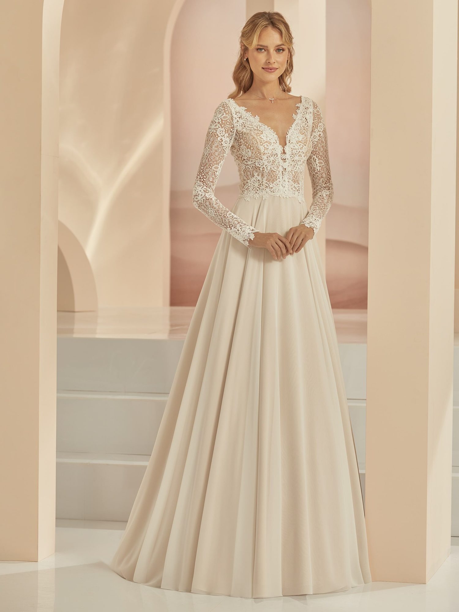 Bianco Evento model Famosa. A-line brudekjole i blonder og chiffon. Kjolen har lange ærmer, dyb v-udskæring foran og bagpå og er både champagnefarvet og ivory.