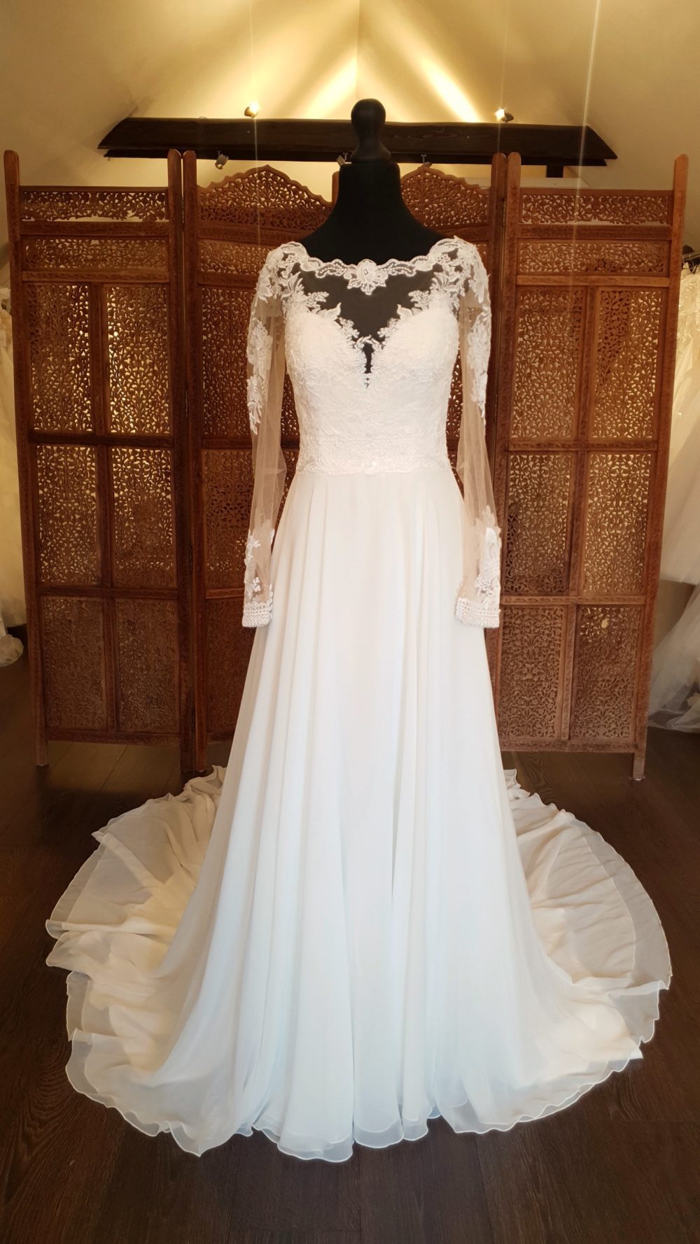 Lillian West. A-line brudekjole med lange transparente ærmer, dekorative blonder på overkroppen, transparent lukket ryg, et skørt i chiffon og et langt slæb.