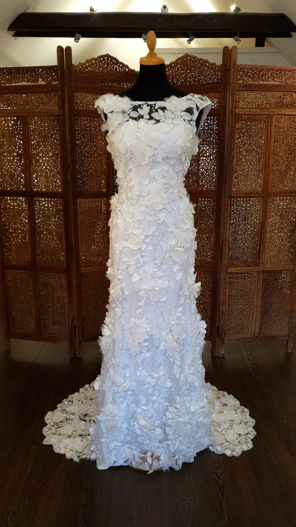 Smuk unik skrædderesyet brudekjole fra Rikke Tudnitz. Den slanke kjole er 2 delt bestående af en glat inderskjole med snøre i ryggen og en blondekjole udenpå fyldt med 3-D blomster. Kjolen har bådudskæring, små korte ærmer og et virkelig smukt slæb.