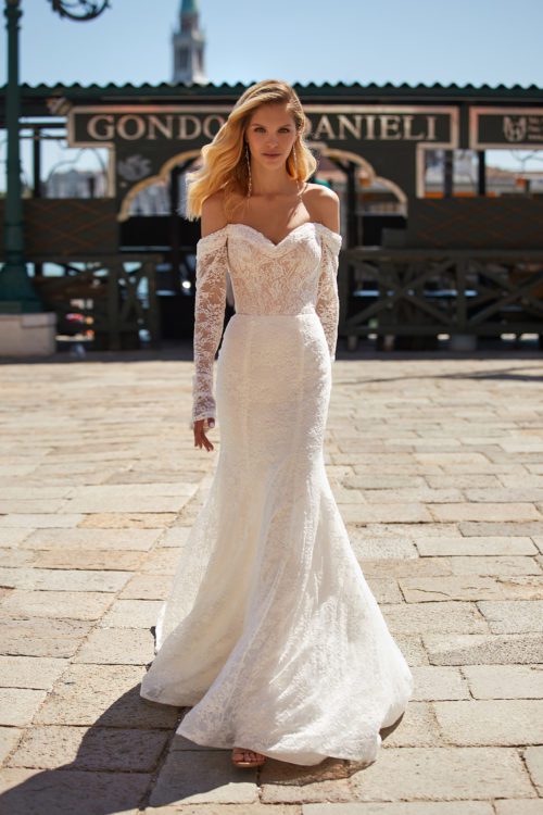 Katy Corso. Model Aster. Slank helblondekjole med lange ærmer der sidder off-shoulder. Kjolens corsage har hjerteudskæring og snøre i ryggen. Skørtet har langt slæb.