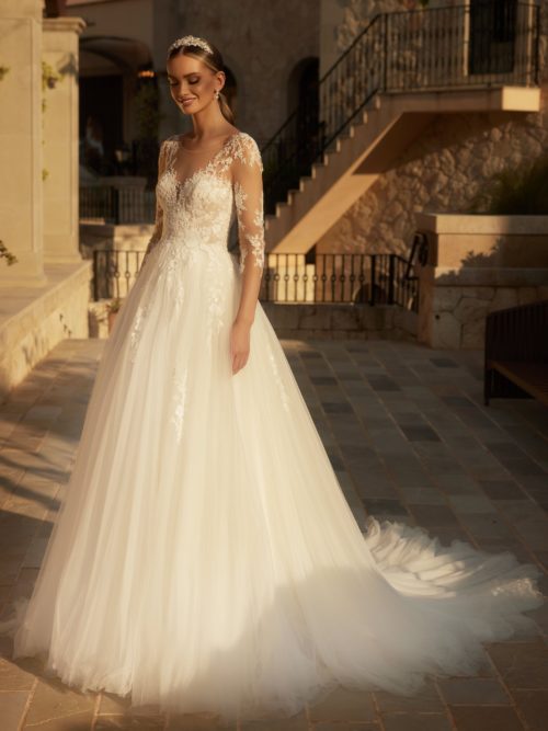 Bianco Evento. Model Carnelian. Prinsessekjole i tyl med 3/ lange transparente ærmer. Kjolen har illusionen af hjerteudskæring og overkroppen har de smukkeste blonder, der er pyntt med perler og pailetter. Ryggen er lukket men meget dekorativ med tatoo-effekt. De smukke blonder fortsætter også ned af skørtet.