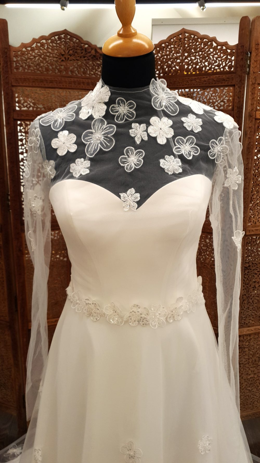 A-line brudekjole bestående af model Harmonia corsagekjole i chiffon fra Bianco Evento og en langærmet kappe i mesh med 3-d blomsterapplikationer. Dertil et blomsterbælte.