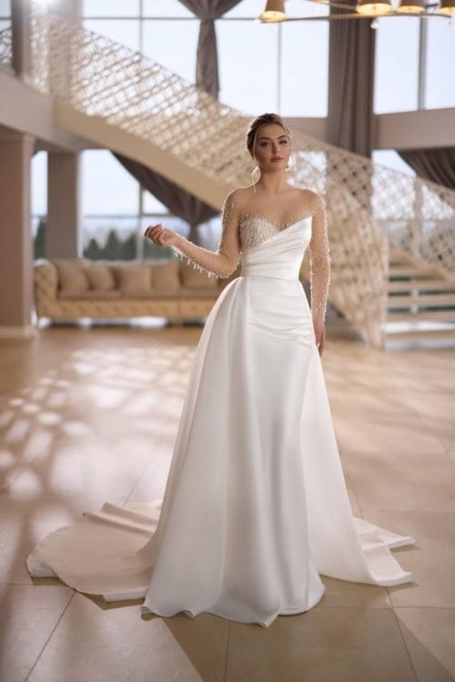 Gellena. Model Holly. Slank brudekjole i satin med unike og smukke perledetaljer. Kjolen har asymmetriske linjer, lange transparente ærmer og et aftageligt slæb i organza.