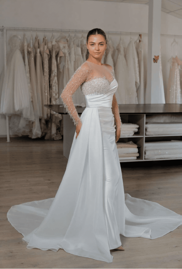 Gellena. Model Holly. Slank brudekjole i satin med unike og smukke perledetaljer. Kjolen har asymmetriske linjer, lange transparente ærmer og et aftageligt slæb i organza.