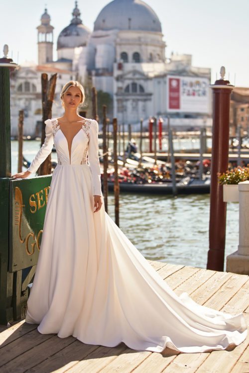 Katy Corso. Model Amber. Smuk, klassisk, elegant og moderne A-line brudekjole i den blødeste satin. Kjolen har lange satinærmer, dyb smal udskæring foran og en smuk dyb v-udskæring på ryggen. På siden af kroppen er der kig blondestof som er transparent og breder sig op til skuldersømmen. Skørtet har masser af vidde, et langt slæb og lommer.
