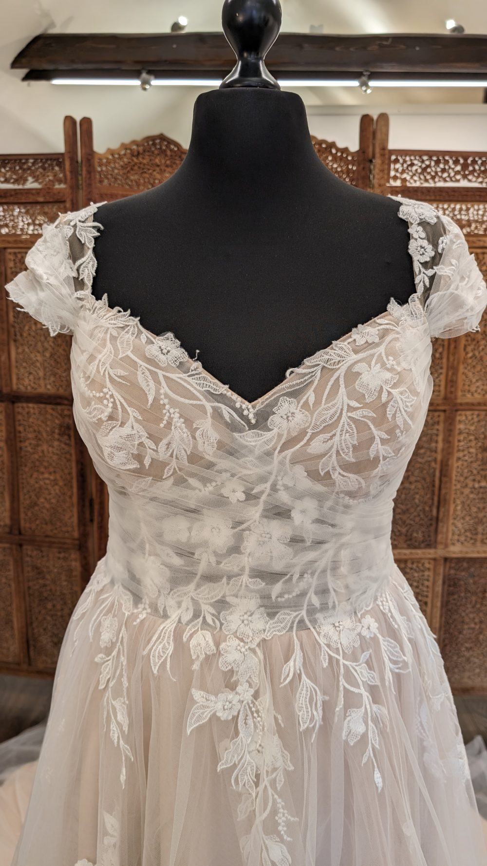 Lillian West. Smuk og romantisk A-line brudekjole. Brudekjolen er beigh med ivory tyl og blondeapplikationer. Den er let transparent i overkroppen, har små korte ærmer, stofbetrukne knapper i ryggen og et lang slæb.