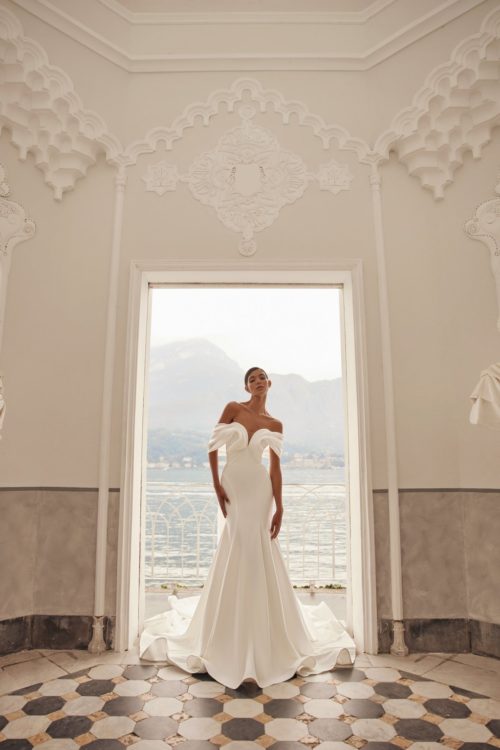 Olya Mak. Model Charmy. Smuk og elegant slank brudekjole i crepe med draperet organza der fra talje og hen over barmen falder ud i bløde off-shoulder ærmer og til sidst danner vinger der folder sig ud på kjolens slæb.