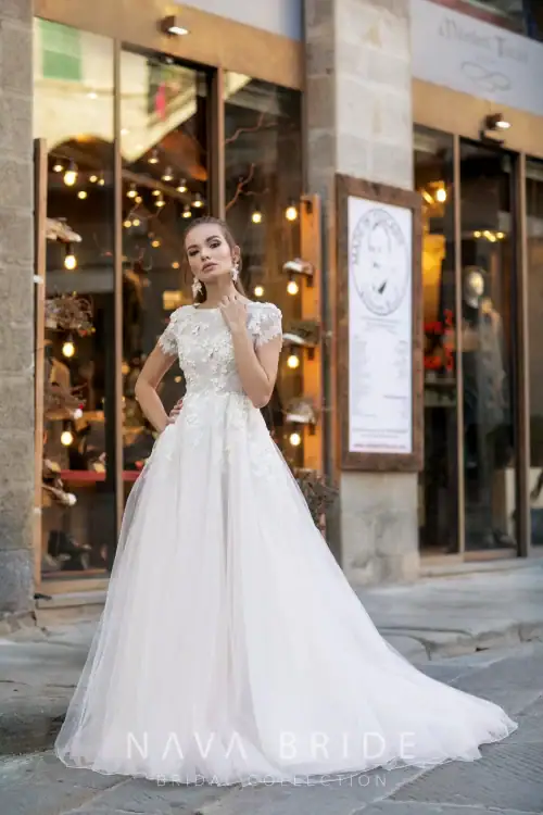 Nava Bride. Model Konstantia. Smuk A-line brudekjole i tyl med høj halsudskæring, små korte ærmer og lukket ryg. Overkroppen har blonder der er dekoreret med små blomsterapplikationer, som fader ned på skørtet.