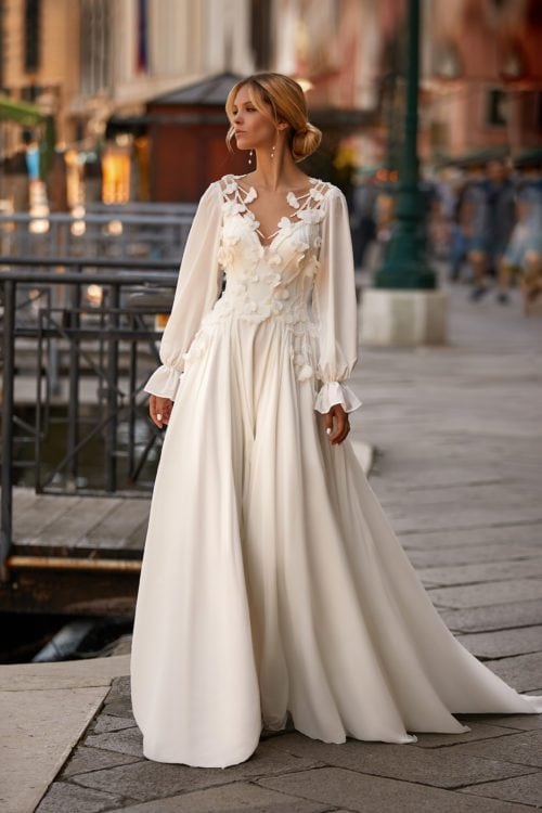 Katy Corso. Model Paula. Smuk og romantisk A-line brudekjole i chiffon med lange ballonærmer og 3-D blomsterapplikationer på overkroppen, som fader ned på skørtet. Kjolen har illusionen af v-udskæring foran og ryggen er lukket med nude mesch.