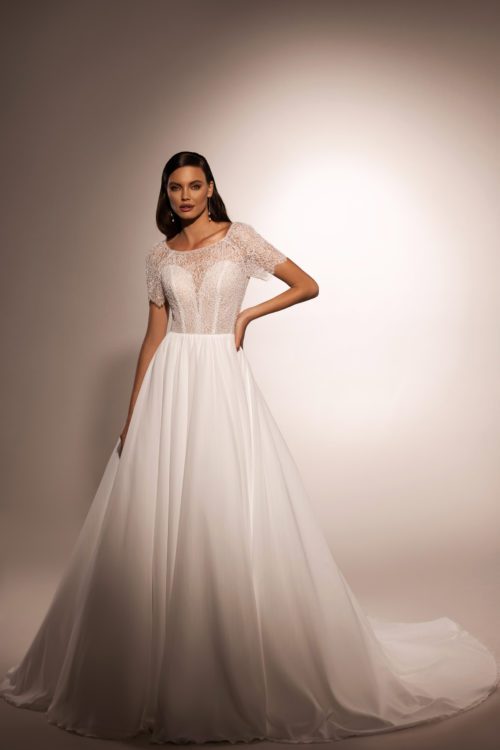 Katy Corso. Smuk og romantisk A-line brudekjole med et enkelt skørt i chiffon. Overkroppens hjerteudskåret corsage er dækket med en fin blonder der danner båduskæring og små korte ærmer. Hele blonden er pyntet med perler og pailetter.