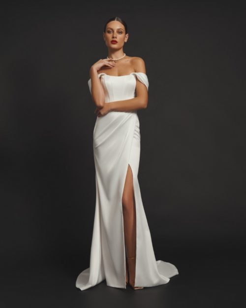 Olya Mak. Model Tobby. Enkel og elegant brudekjole i satin. Kjolen corsage er bådudskåret og har off-sholder stropper, samt snøre i ryggen. Skøret har slå-om effekt, med draperinger der går op til taljen og en lang lidt åben slids ved venstre ben. Lille slæb.