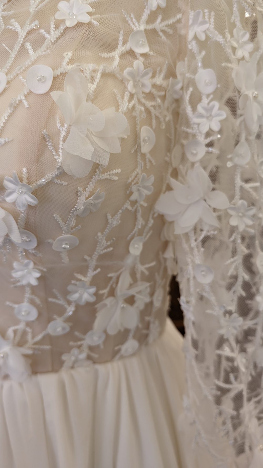 The Wedding Dress. Model Ethna. A-line brudekjole i chiffon. Kjolens overkrop består af en nude corsage beklædt med mesh der danner høj rund halsudskæring, lukker ryggen af og giver den lange ærmer. Overkroppen er dekoreret med masser af 3D blomster.