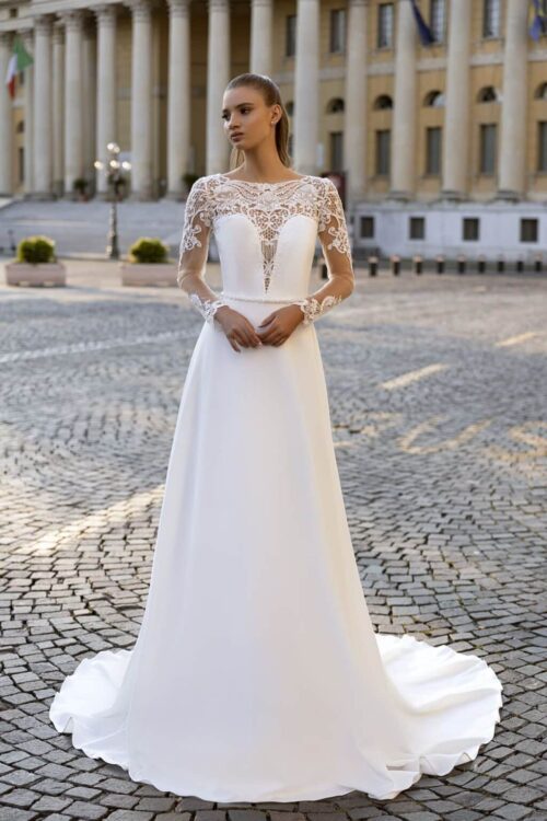 Bella by Eglantine Creation. Model BEL22431. Smuk langærmet kjole i crepe. Kjolen har en corsage med dyb cavalergang der er dækket af en meget smuk bådudskåret blonde, der går ned i v-udskæring på ryggen. Kjolen har stopbetrukne knapper i ryggen.