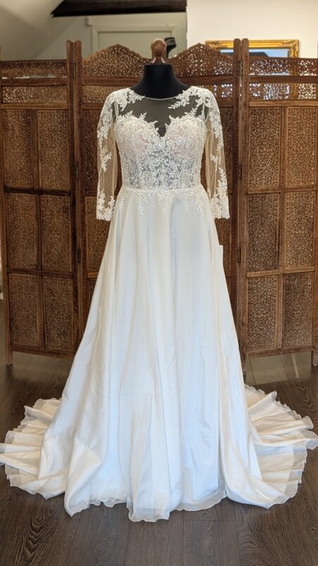 White One. Model Bolingo. Smuk og dekorativ A-line brudekjole med et let skørt i organza. Kjolen har hjerteudskæring indenunder en nude mesch, som danner rund halsudskæring og 3/4 lange ærmer. Ryggen er lukket med stofbetrukne knapper og hele overkroppen er dekoreret med de smukkeste blonder har har transparente områder.