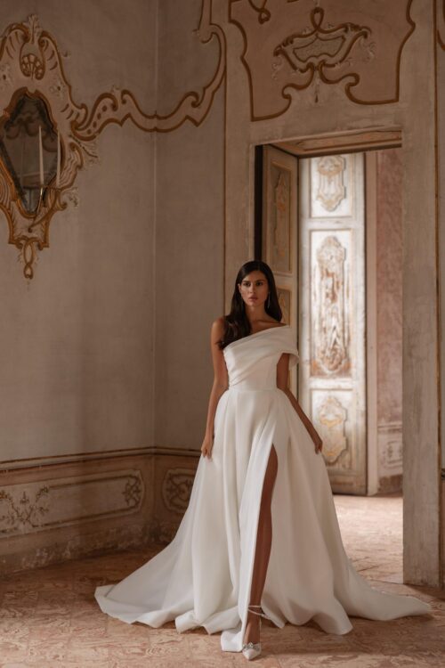 Ivory Bridal Fashion. Model Clarins. Smuk minimalistisk prinsessekjole i organza. Kjolen er elegant og feminin. Den har et asymmetrisk look med sin brede off-shoulder ærme på den ene arm og en stor smuk tvistet sløjfe bagpå. Kjolen har yderligere høj slids i skørtet foran og et lang slæb.