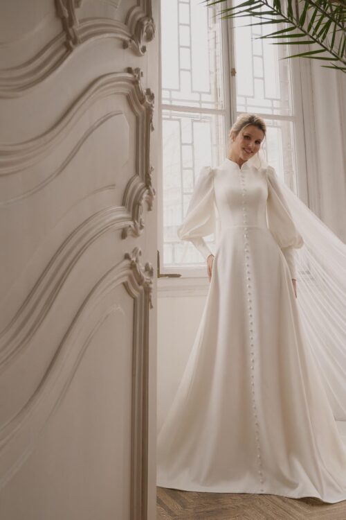 Ivory Bridal Fashion. Model Amalia. Smuk minimalistisk A-line brudekjole i satin. Kjolen har lange ærmer med balloneffekt på overarmen, høj halsudksæring med lukket ryg og kinakrave. Som en meget elegant detalje har den stofbetrukne knapper hele vejen ned midt foran fra hals til gulv.