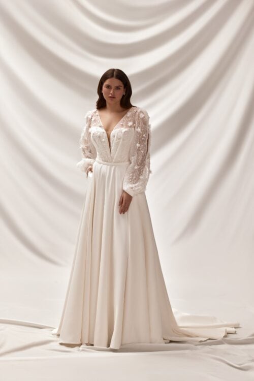 Melory. Model Octavia. Smuk A-line brudekjole i satin med hjerteudskæring og dyb kavalergang. Kjolen overkrop er dækket med en Ivory mech med lange ballanærmer. Meschen er dekoreret med perler, pailetter og 3-D blomster. Den er drapperet forna og danner v-udskæring både foran og bagpå.