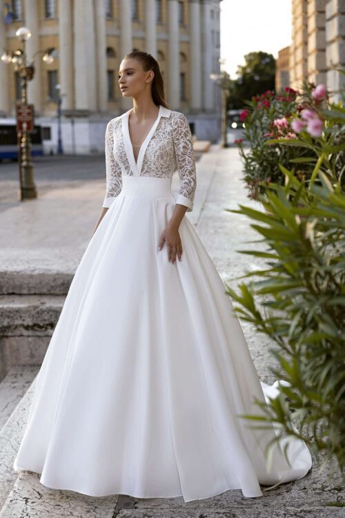Bella by Eglantine. Model BEL22410. A-line brudekjole i satin med en overkrop som skiller sig ud fra mængden. Kjolen har blonder, 3/4 lange ærmer, dyb v-udskæring og lukket ryg. Satin kanterne i udskæringen danner også en lille krave i nakken og fine afslutninger på ærmerne. Skørtet har et bredt bærestykke i taljen og store læg i skørtet som giver det masser af volumen.