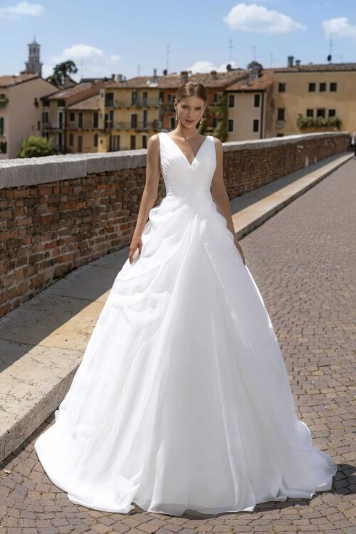 Bella by Eglantine. Model BEL22432. Klassisk A-line brudekjole i organza med drapperet overkrop og asymmetriske læg i skørtet. Kjolen har brede stroppe, v-udskæring foran og en lidt dybere v-udskæring bagpå.
