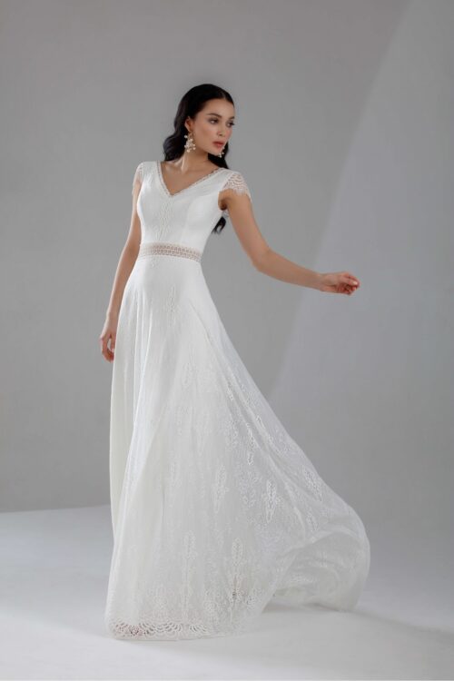 Jarice Bridal. Model Art 24-21. Smuk helblondekjole i en slank A-line. Kjolen har et transparent bånd i taljen, v-udskæring foran og bagpå samt små korte blondeærmer.
