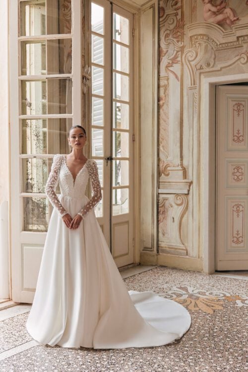 Katy Corso. Model Delara. Smuk A-line brudekjole i satin med lange ærmer og lommer. Kjolen har dyb udskæring foran og er fyldt med små 3-D blomster på både krop og ærmer.