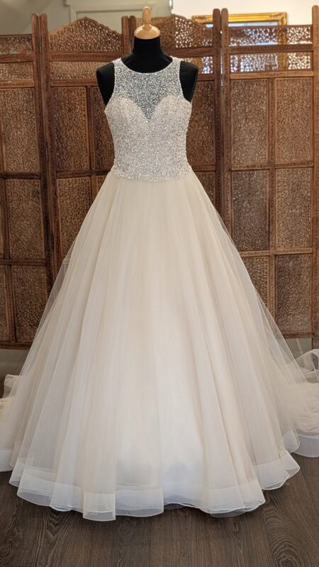 Morilee by Madeline Gardner. Smuk A-line prinsessekjol i tyl med en overkrop fyldt med rhinsten. Kjolen er champaggnefarvet, har høj rund halsudskæring foran med en hjerteudskåret corsage under den pyntede mesh. På ryggen er der et stort key-hole.