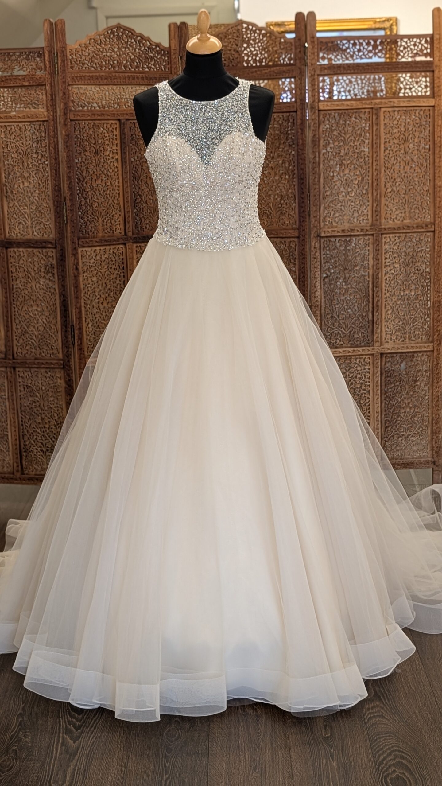 Morilee by Madeline Gardner. Smuk A-line prinsessekjol i tyl med en overkrop fyldt med rhinsten. Kjolen er champaggnefarvet, har høj rund halsudskæring foran med en hjerteudskåret corsage under den pyntede mesh. På ryggen er der et stort key-hole.