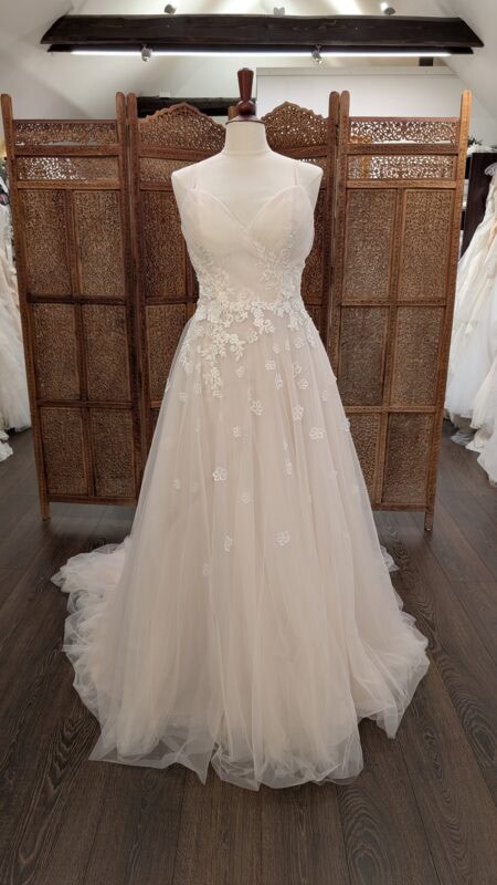 Phoenix Gowns. Model PH0089. Smuk og romantisk A-line brudekjoe i tyl. Kjolens farve er lys sand, overkroppen har v-udskæring, er drapperet og har spagettistropper. Den har smukke blonder, som bevæger sig fra overkroppen og ned på det lette skørt. Den har dyb rygudskæring og et langt slæb.