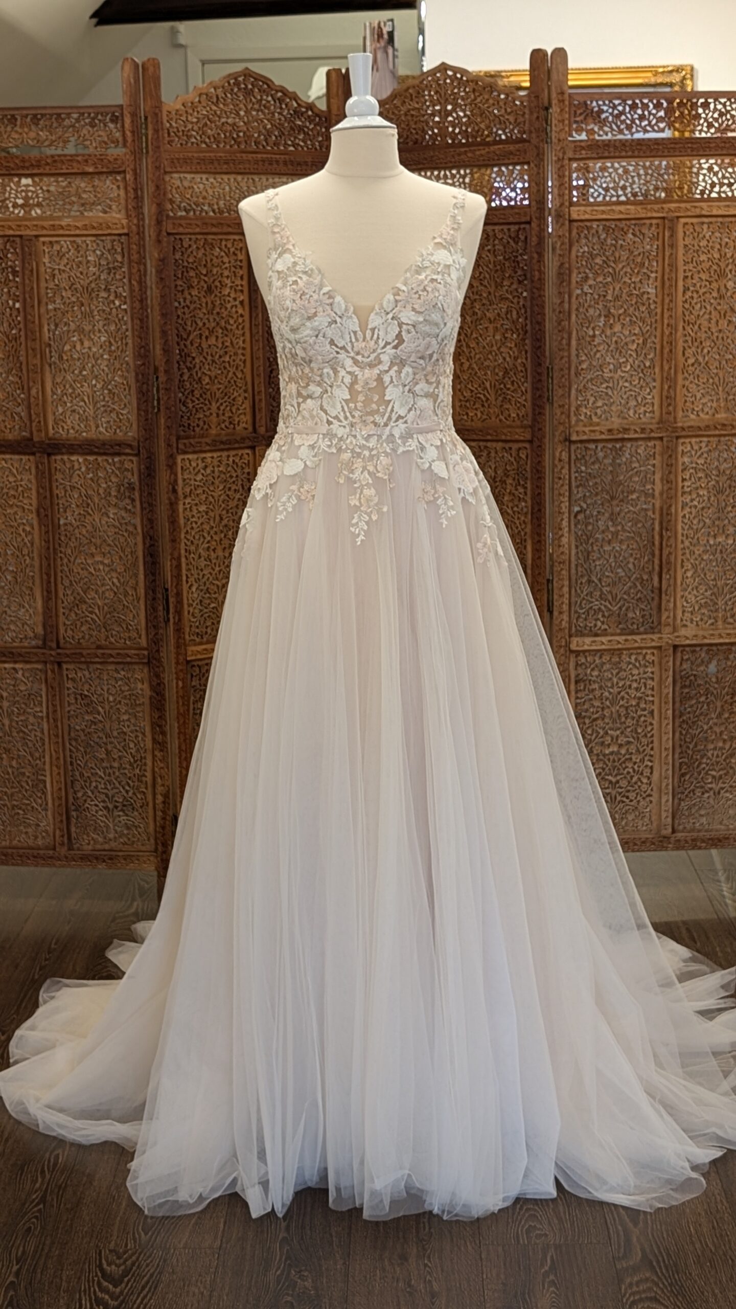 Maggie Sottero. Model Minerva Blush. Smuk og romantisk A-line brudekjole i tyl med masser af blondeapplikationer i sarte farver. Kjolen er blush og har v-udskring foran og dyb v-udskæring på ryggen. Den har et satinbånd omkring taljen, knapper i ryggen og et smukt og dekorativt slæb.