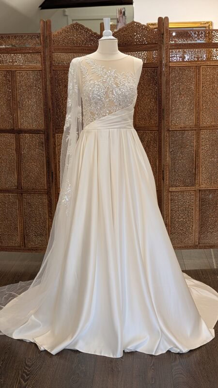 Milan Bride. Smuk A-line brudekjole i satin. Kjolen har asymmetriske deltajer via drappringere i taljen og med et enkelt "kappe"-ærme der fra skulderen falder ud over armen og fortsætter med ud på slæbet. Overkroppen er transperent og dekorert med blonder, perler og pailetter. Ryggen går ned i en dyb åben v-udskæring.