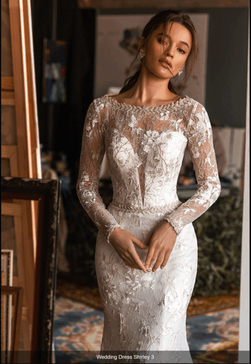 Jasmin Empire. Model Shirley. En smuk og dekorativ slank og langærmet brudekjole med kobbertråd og broderinger. Kjolen har illusionen af dyb kavalergang, men har høj bådudskæring . Ryggen er lukket men helt transparent og giver illusionen af dyb udskæring.