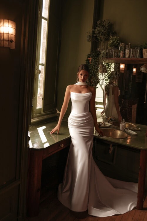 Katy Corso. Model Nanny. Elegant slank brudekjole i satin. Kjolen er uden stropper, har bådudskæring med draperinger over barmen og omkring hofter og mave.