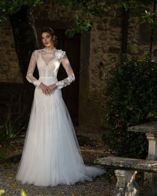 Hadassa. Model Evangeline. Let og elegant brudekjole i tyl. Kjollen har en sart blonde som dækker en v-udskåret corsage med tynde stropper. Blonden danner lange ærmer og høj hals. Der er knapper i ryggen og på ærmer og den ene skulder er der påsyet smukke 3-d blomster. Blonden bevæger sig ned over hoften og går derefter ud i et fyldigt tylskørt med slæb.