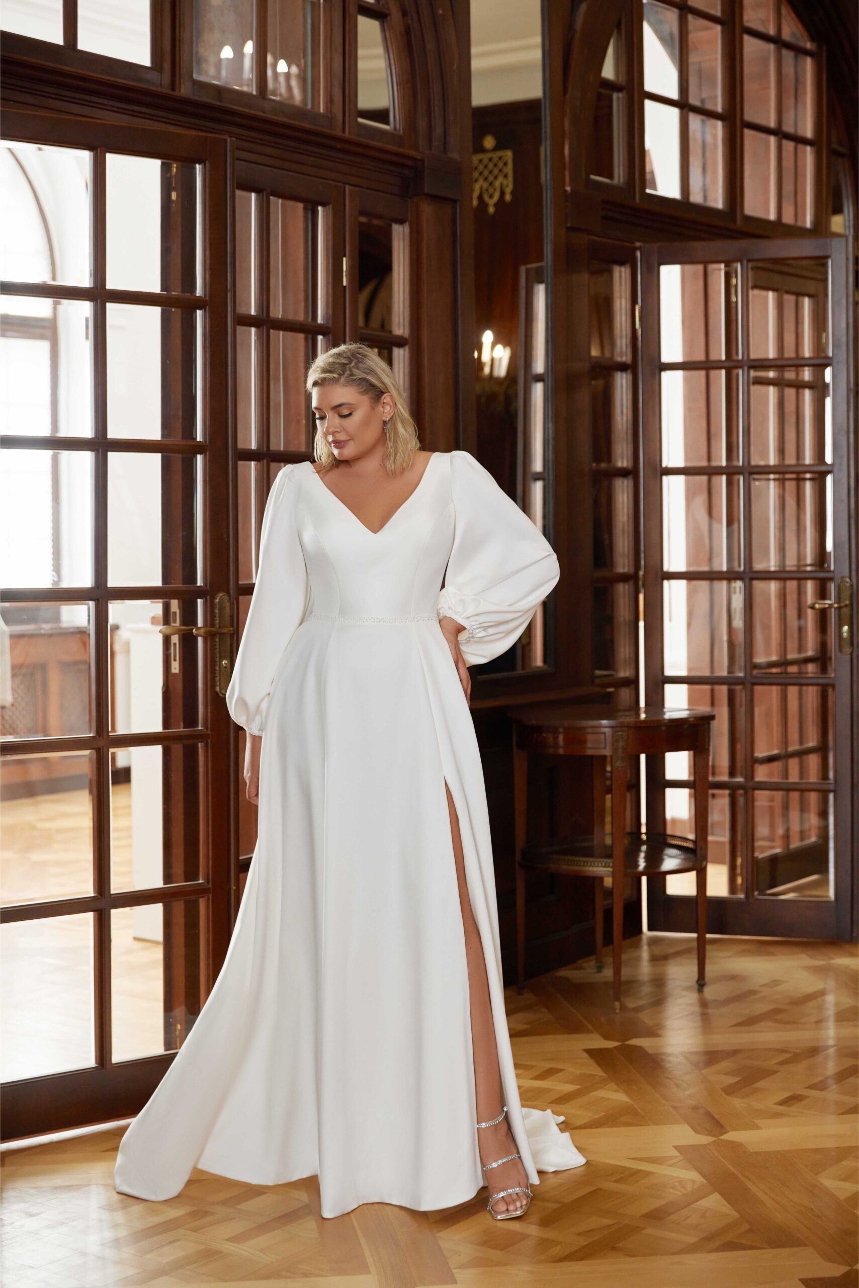 Kaya Nova. Model Soha. Elegant og smuk minimalistisk A-line brudekjole i crepe. Kjolen har lange ballonærmer, v-udskæring både foran og bagpå, samt høj slids i skørtet ved venstre ben.