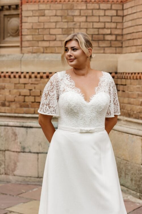 Kaya Nova. Model Rabia. Smuk A-line brudekjole med blonder på overkroppen og satinskørt. Kjolen har en hjerteudskåret corsage under blonden deer danner v-udskæring både foran og bagpå. Den har korte flagermusmærmer og et påsyet bånd med sløjfe i taljen.