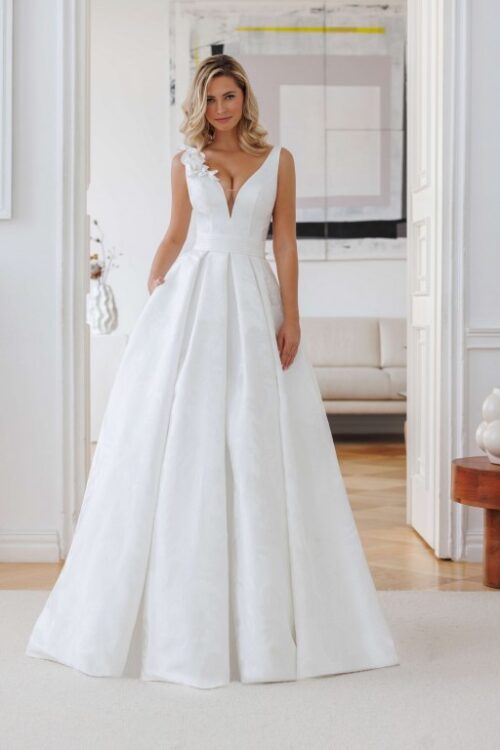 Jarice Bridal. Model Ashley. Smuk A-line brudekjole i Jaquard stof med blomsterprint. Kjolen har brede stropper med 3-D blomster på højre skulder. V-udskæring både foran og bagpå, store læg i skørtet og lommer.
