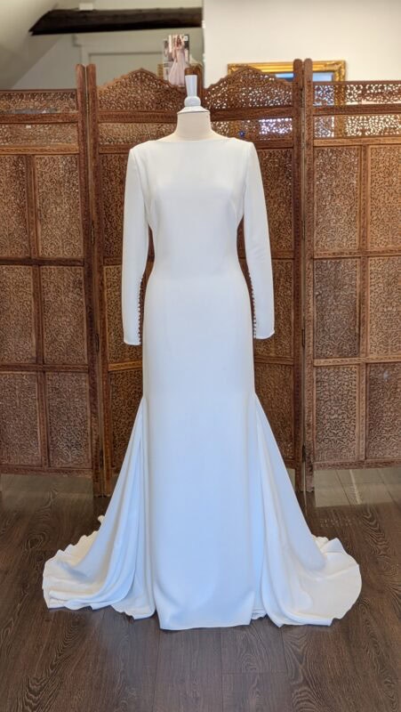 Pronovias. Model Leona. Slank minimalistisk og elegant brudekjole i crepe med lange ærmer og dyb udskæring på ryggen. Den har bådudskæring foran og smukke stofbetrukne knapper i ryggen og i ærmerne.