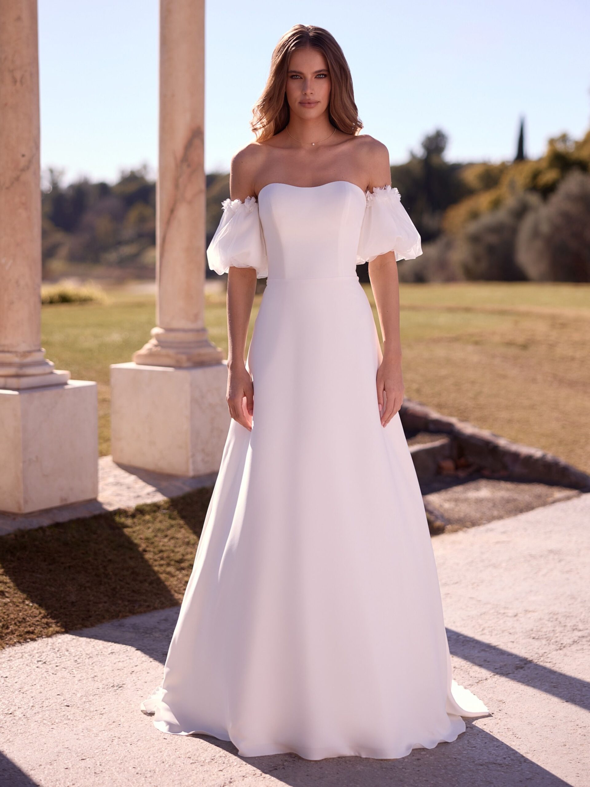 Bianco Evento. Model Paulette. Romantisk og enkel brudekjole i strech crepe. Kjolen er stropløs og har hjerteudskæring. De små ballonærmer er aftagelige og har fine 3-d blomster i kanten.