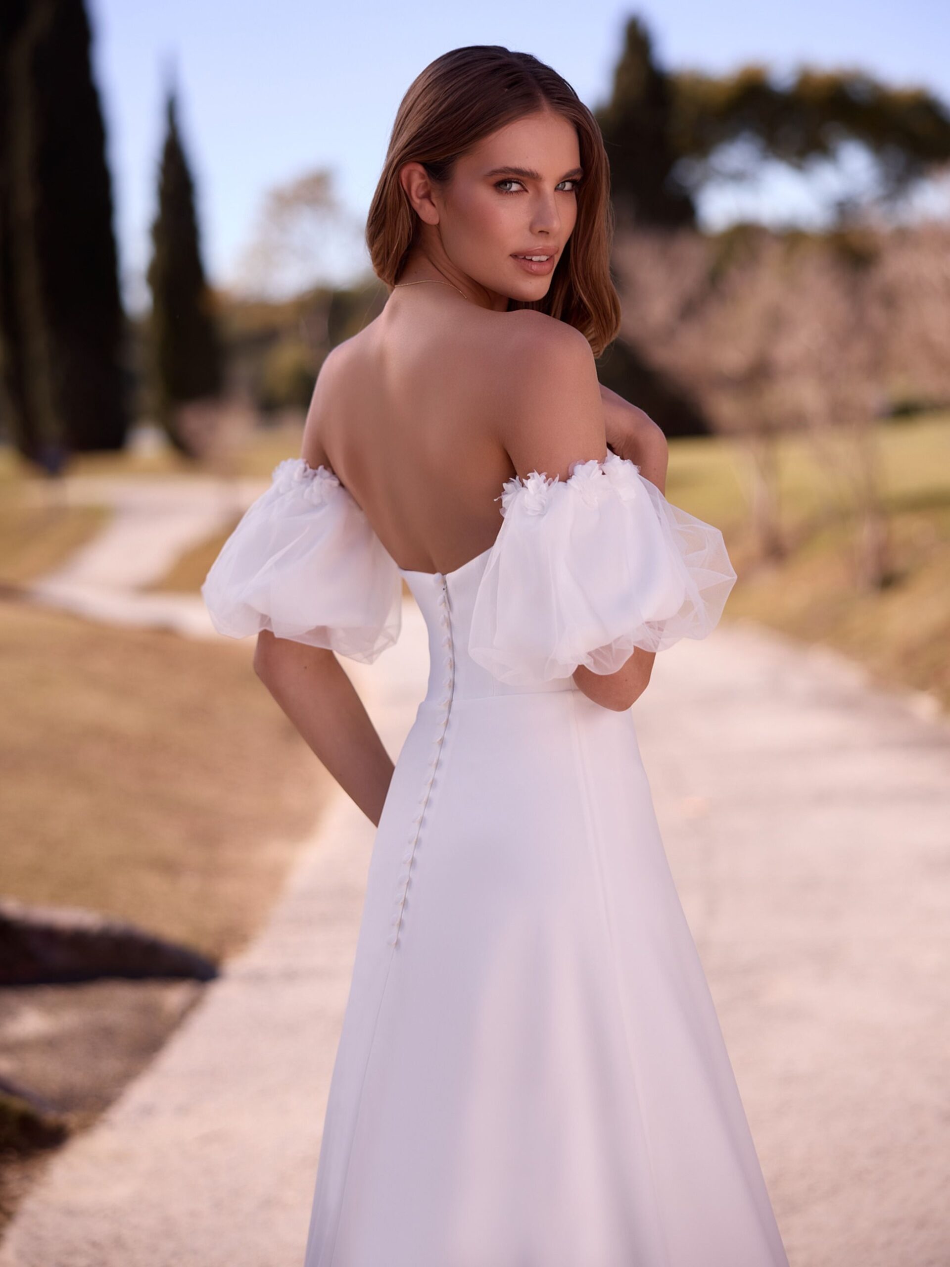 Bianco Evento. Model Paulette. Romantisk og enkel brudekjole i strech crepe. Kjolen er stropløs og har hjerteudskæring. De små ballonærmer er aftagelige og har fine 3-d blomster i kanten.