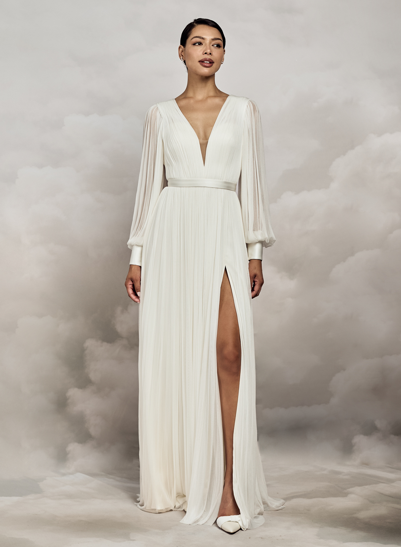 Catherine Deane. Model Levi. Let og elegant langærmet A-line brudekjole i 100 % silketyl. Kjolen har dyb v-udkskæring både foran og bagpå. Ballonærmerne er transparente og afslutes med en smuk bred manchet der lukkes med stofbetrukne ærmer. Kjolen har et påsyet glat bånd i taljen, høj slids i skørtet og et lille slæb.