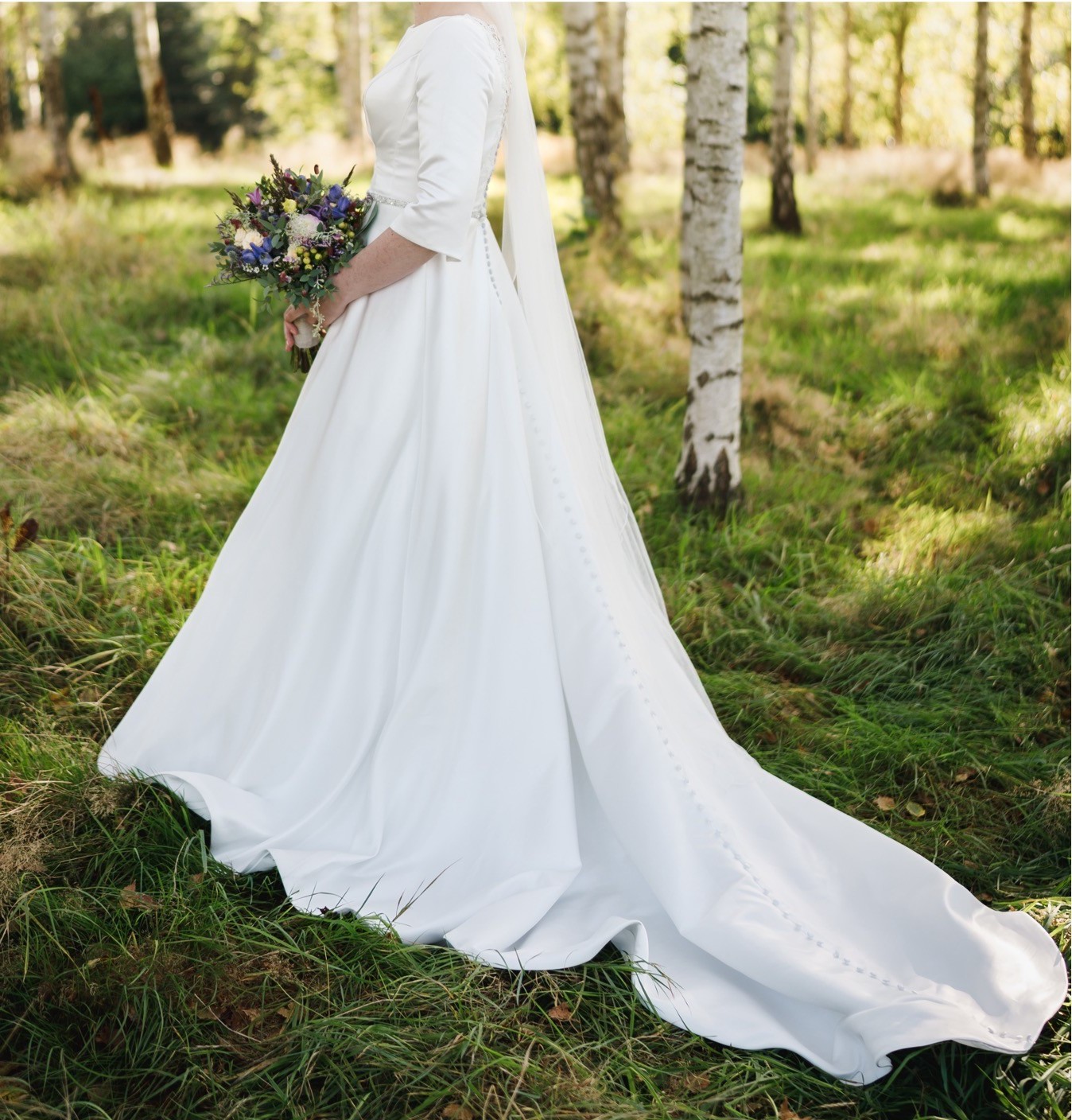 Phoenix Gowns. Model PH0033. Klassisk og enkel brudekjole i glat satin. Kjolen har små fine deltaljer som påsyet perlebælte i taljen, bådudskæring med en smal v-udskæring foran. Dyb trantsparent ryg med dekoratie blonder, små korte ærmer og stofbetrukne knapper hele vejen ned på slæbet.