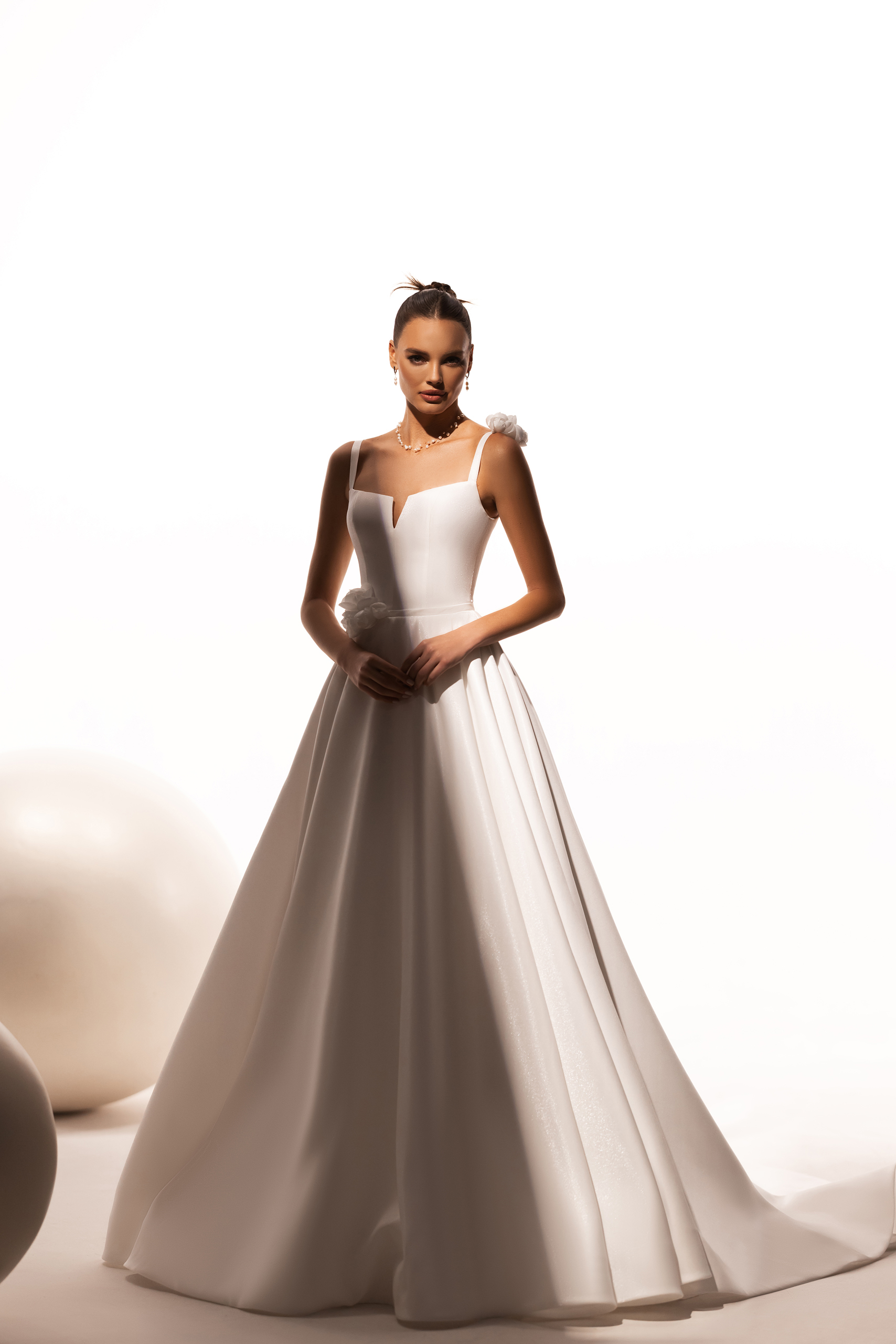 Katy Corso. Model Lilibet. Smuk og enkel A-line kjole med smukke 3-d blomster detaljer. Kjolen har stropper, lige skæring over barmen med en lille v-udskæring. Blomsterne er plaseret på den ene skulder og i modsatte side af taljen. Ryggen har en dyb forkatet udskæring. Stoffet er kraftigt og falder utrolig flot. Det helt særlige er dens perlemors shine som er diskret men utrolig smukt.