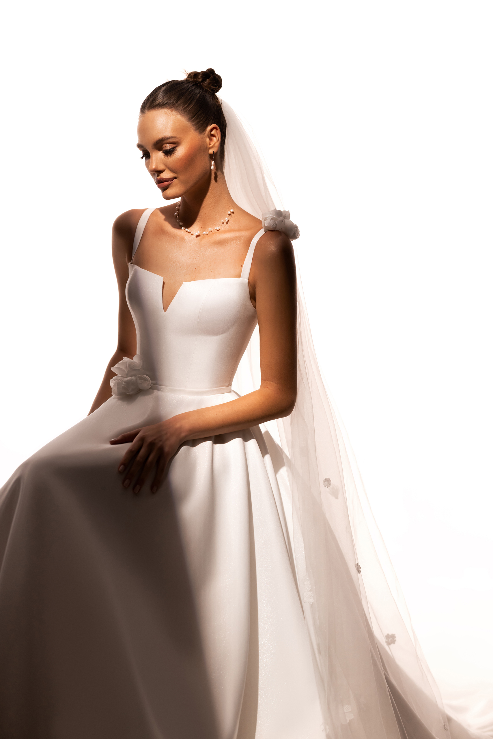 Katy Corso. Model Lilibet. Smuk og enkel A-line kjole med smukke 3-d blomster detaljer. Kjolen har stropper, lige skæring over barmen med en lille v-udskæring. Blomsterne er plaseret på den ene skulder og i modsatte side af taljen. Ryggen har en dyb forkatet udskæring. Stoffet er kraftigt og falder utrolig flot. Det helt særlige er dens perlemors shine som er diskret men utrolig smukt.