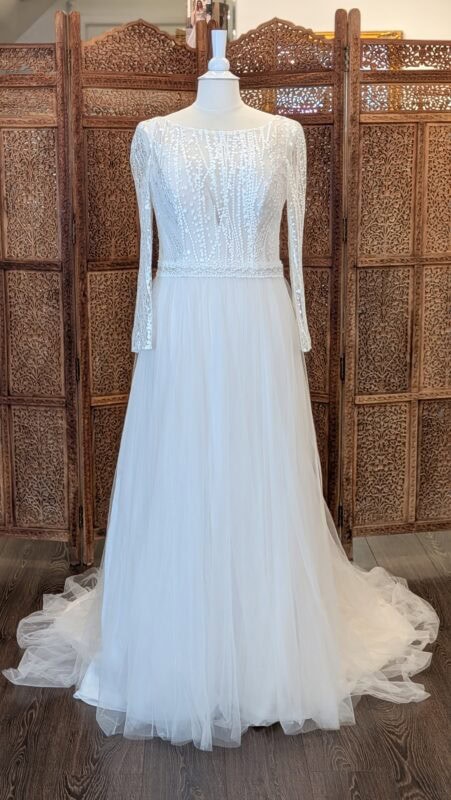 Le Papillon. Model Elena. Smuk A-line brudekjole i tyl. Kjolen har bådudskæring foran men med en nude corsage med dyb udskæring indenunder den Ivory mesh. Den har lange ærmer og en dyb åben ryg. Meshen er broderet med lodrette linjer og prikker. Kjolen har et påsyet bånd i taljen med perler.