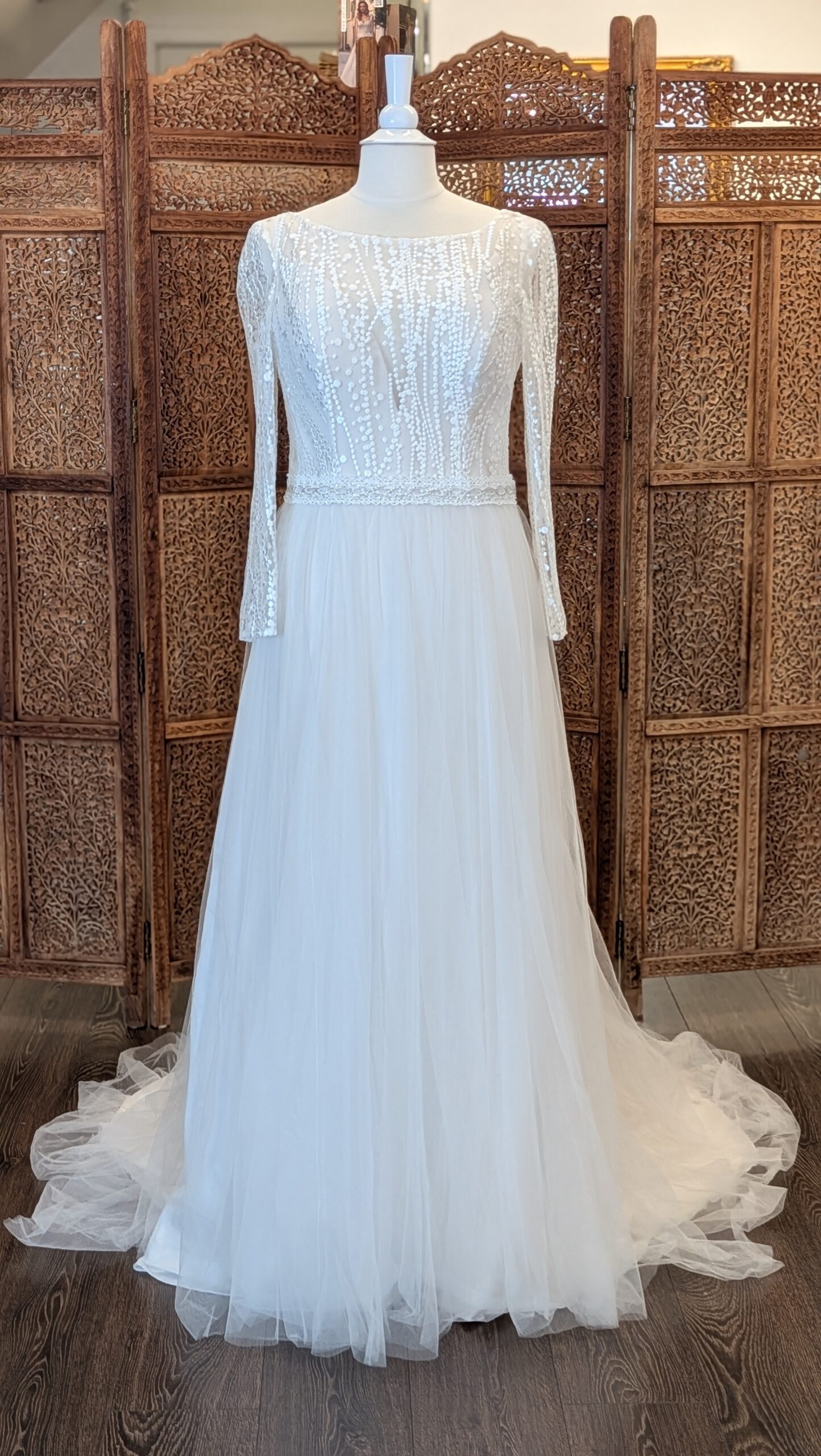 Le Papillon. Model Elena. Smuk A-line brudekjole i tyl. Kjolen har bådudskæring foran men med en nude corsage med dyb udskæring indenunder den Ivory mesh. Den har lange ærmer og en dyb åben ryg. Meshen er broderet med lodrette linjer og prikker. Kjolen har et påsyet bånd i taljen med perler.
