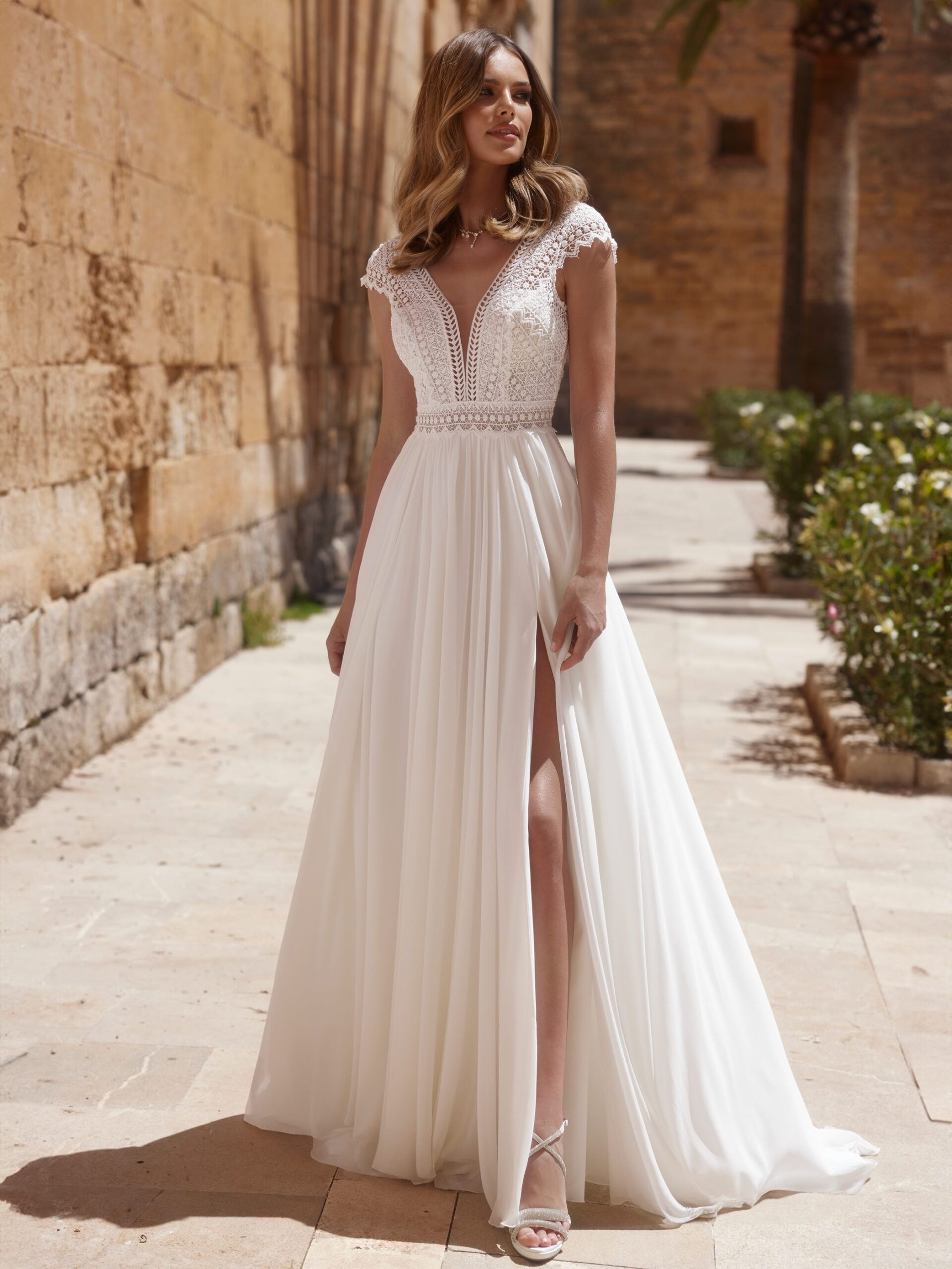 Bianco Evento. Model Warys. Smuk boheme kjole i chiffon. Kjolen har smukke grafiske blonder i overkroppen med dyb v-udskæring både foran og bagpå og små korte ærmer. Skørtet har høj slids og mellemlangtt slæb.