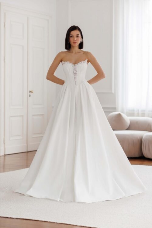 Jarice Bridal. Model Art 26/23. Smuk enkel og stropløs prinsessekjole i satin. Kjolens corsage er hjertudskåret, har synlige stivere og en dyb kavalergang. Den er yderligere forsynes med skåle der titter op over udskæringen der er pyntet med 3-D blomstter og perler. Pynten fortsætter ned af kavalergangen og sidder også rundt i hele taljeskæringen. Skørtet har store læg og slæb.