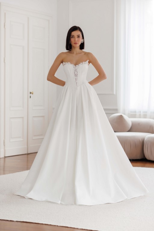 Jarice Bridal. Model Art 26/23. Smuk enkel og stropløs prinsessekjole i satin. Kjolens corsage er hjertudskåret, har synlige stivere og en dyb kavalergang. Den er yderligere forsynes med skåle der titter op over udskæringen der er pyntet med 3-D blomstter og perler. Pynten fortsætter ned af kavalergangen og sidder også rundt i hele taljeskæringen. Skørtet har store læg og slæb.