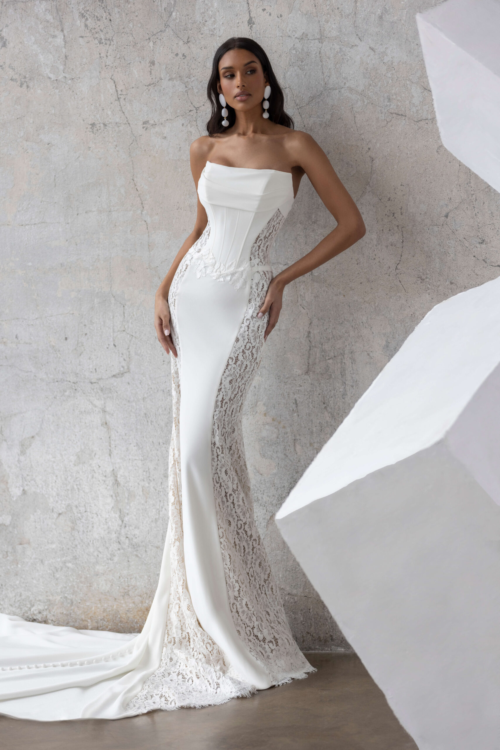 Jarice Bridal. Model Fabienne. Smuk og elegant slank brudekjole. Kjolen er stropløs, har lige skærring over barmen og har detaljer som lette draperinger over barmen, lodrettet stivere i overkroppen og blonder der er dekorert med perler og pailetter i taljen. Det unike ved denne kjole er helt klart de brede blondepartier, som følger fra overkroppen og hele vejen ud på slæbet og fremhæver figuren, på smukkeste vis.